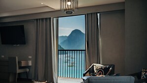 A modern living room with a large window, a flat-screen TV, a dining area, and a balcony with a view of mountains.