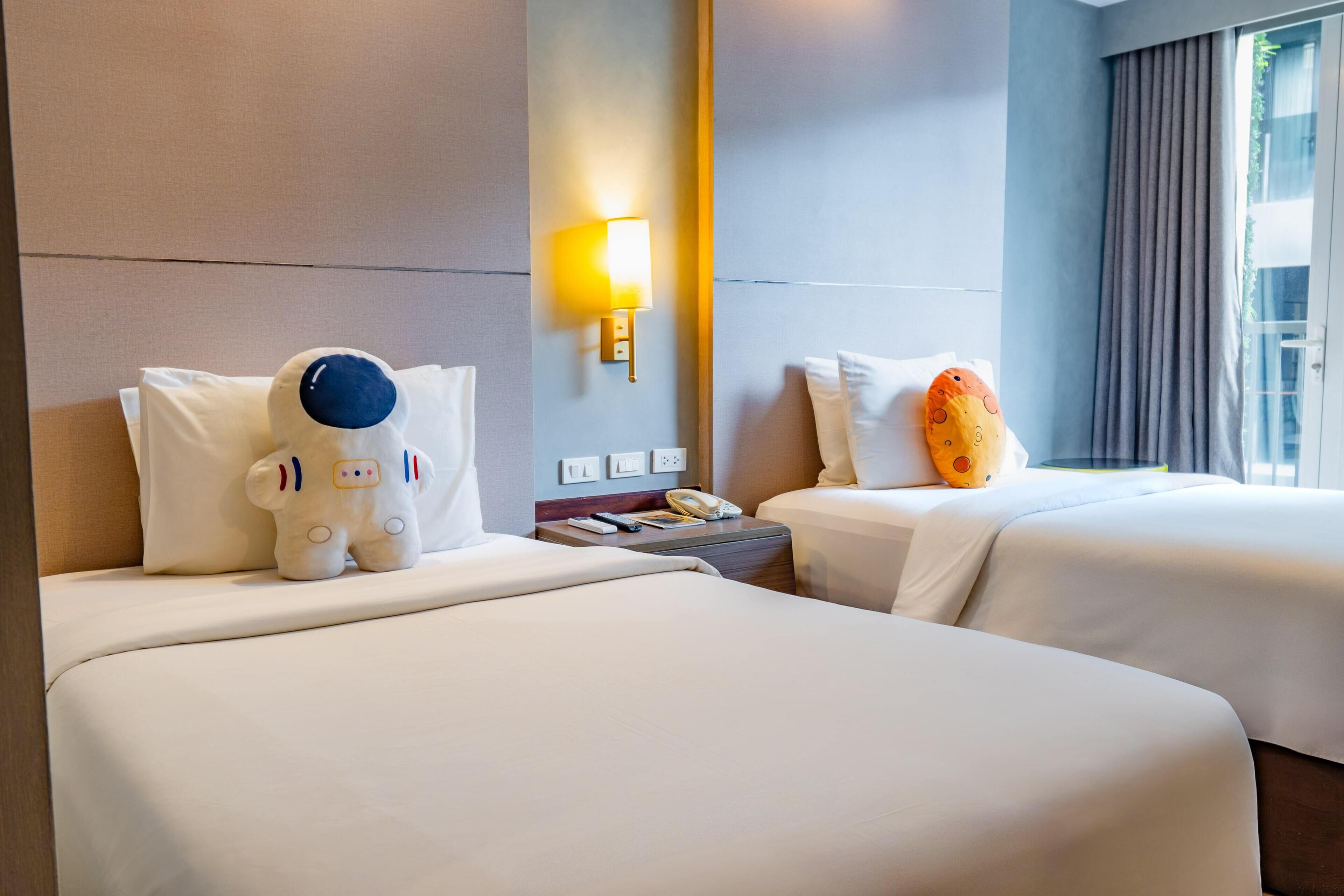Habitación de hotel con dos camas, una mesita de noche con lámpara y una ventana con cortinas.