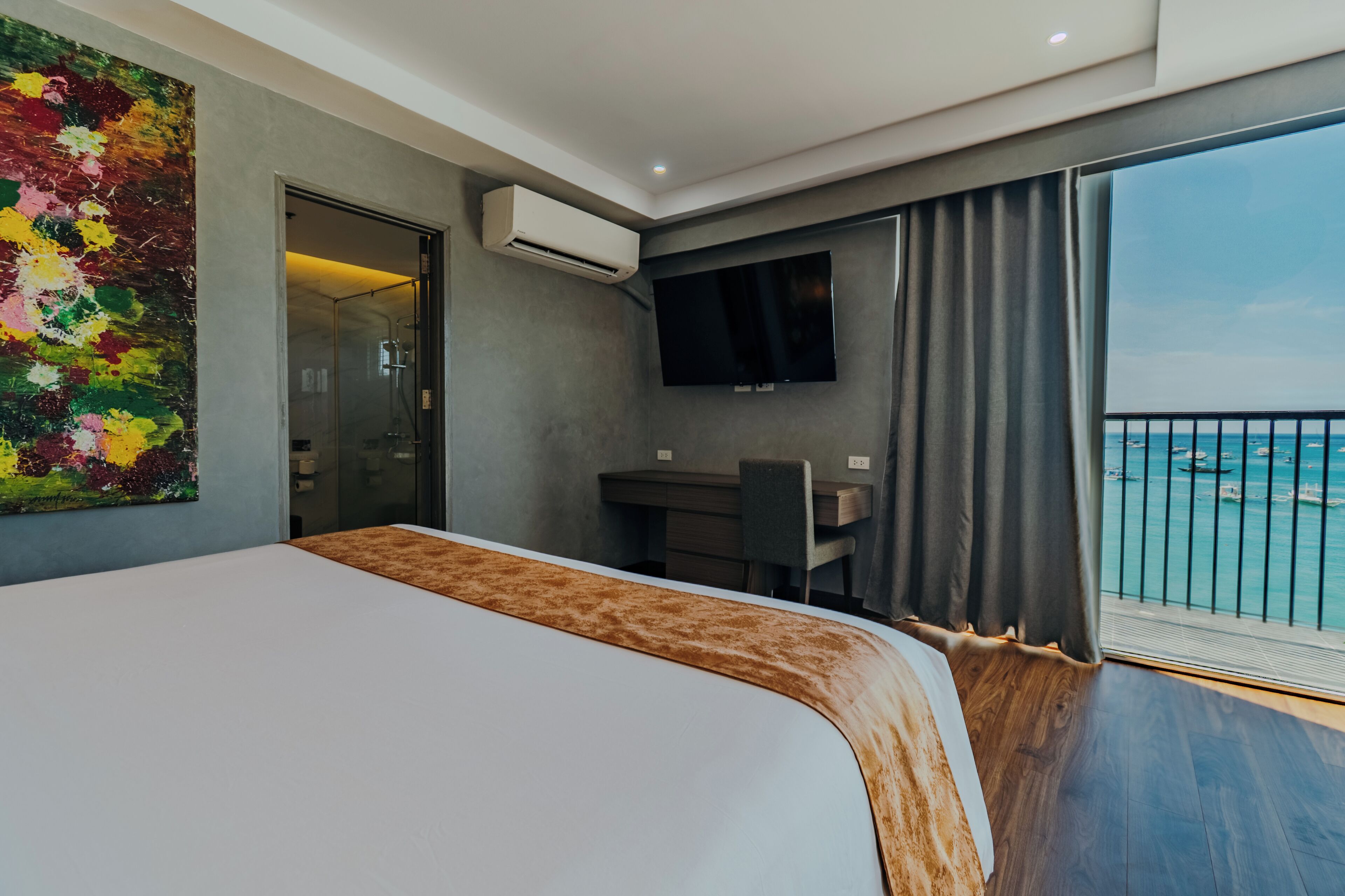 A modern hotel room with a large bed, a desk, a TV, and a balcony with a view of the sea.