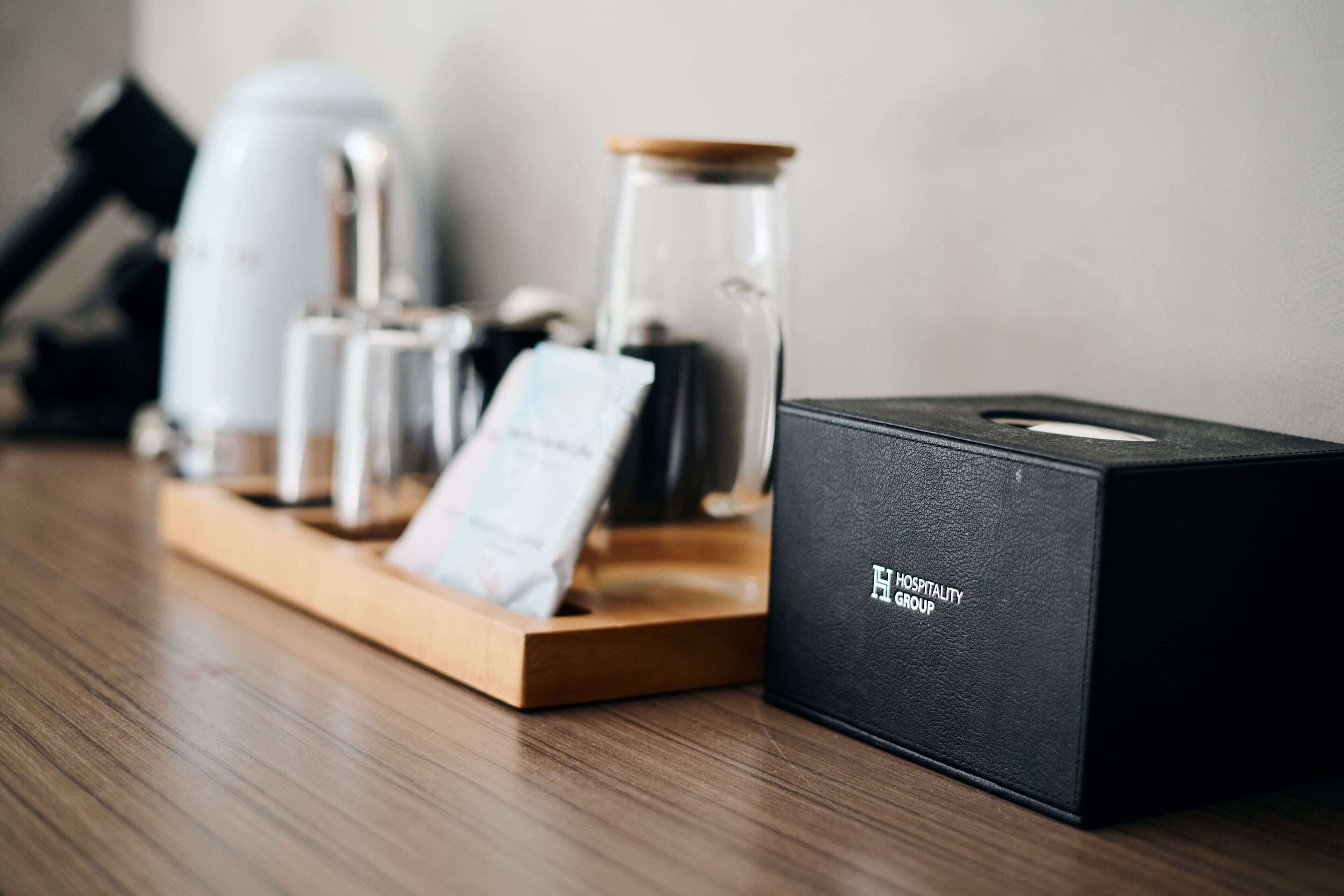 Una scatola nera con la scritta 'HOSPITALITY GROUP' su un vassoio di legno, insieme a una macchina da caffè, un barattolo di vetro e un biglietto.