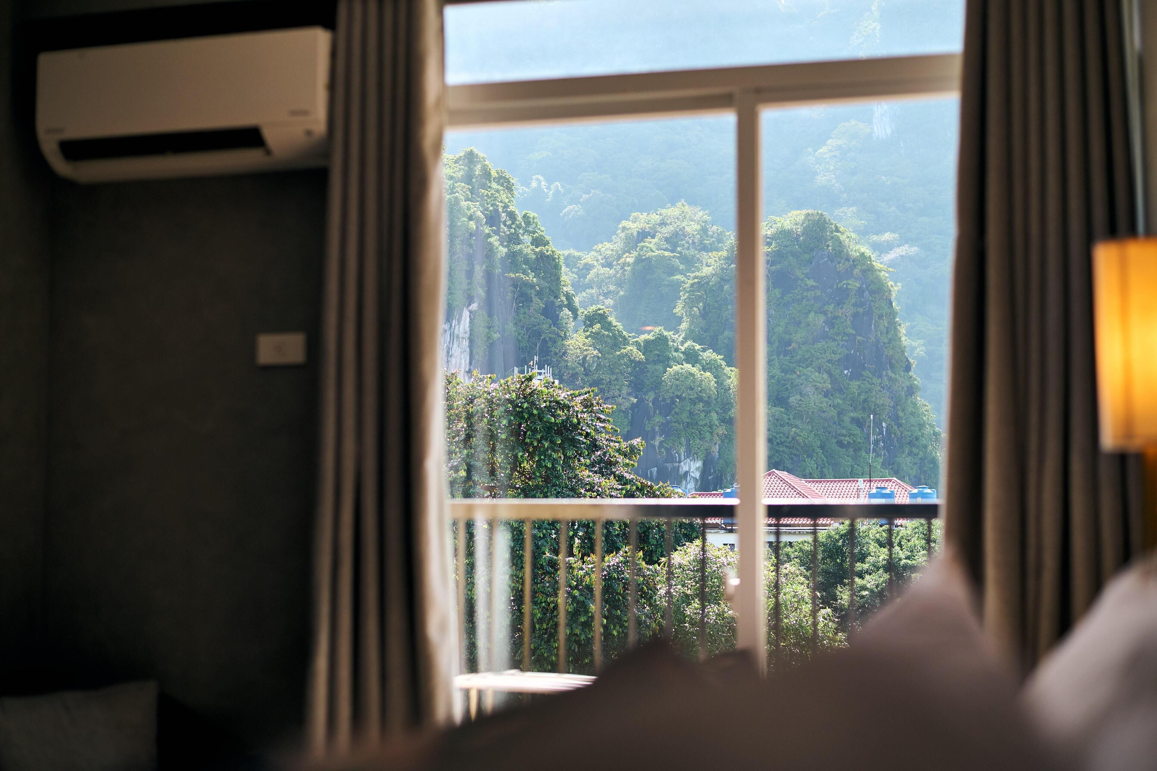 A room with a window overlooking a lush green landscape, a balcony with a railing, and an air conditioning unit on the wall.