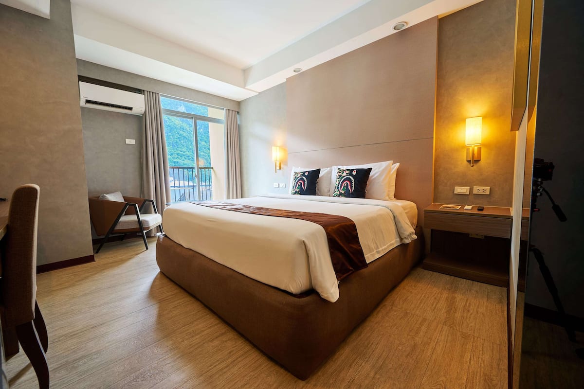 A hotel room with a large bed, a desk, a chair, a balcony, and a wall-mounted lamp.
