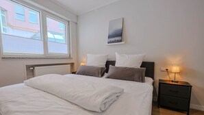 Ein ordentlich bezogenes Bett mit weißen Laken, ein Nachttisch mit Lampe und ein Bild an der Wand.