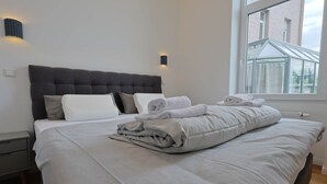 Een net opgemaakt bed met opgevouwen handdoeken, een grijs gestoffeerd hoofdeinde en een raam met uitzicht op een bakstenen gebouw.