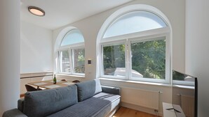 A modern living room with a grey sofa, a dining table, and large arched windows offering a view of trees. - Deluxe Apartment No.03 with 2 bedrooms (Badenweiler)