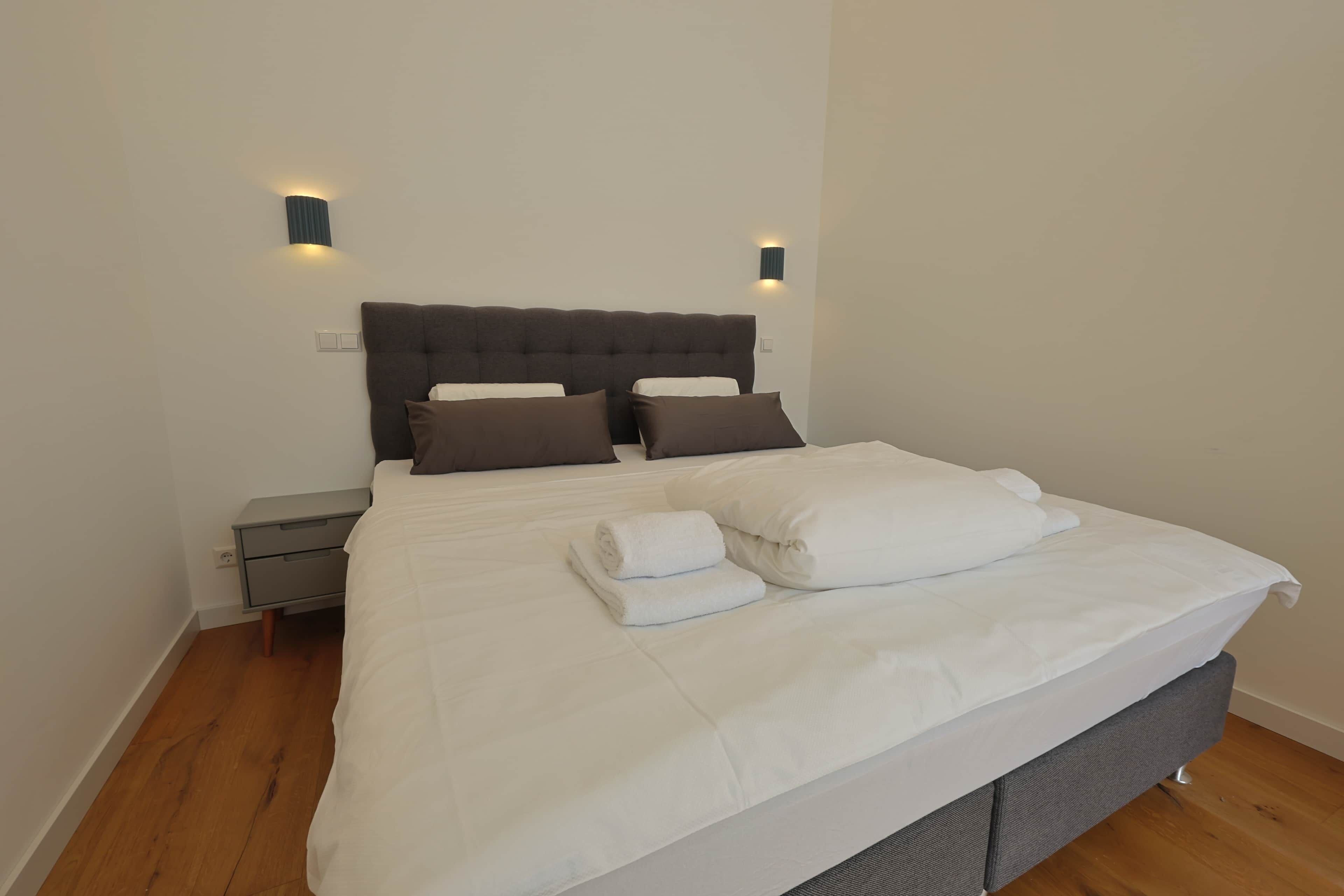 Ein ordentlich bezogenes Bett mit weißen Leinen und braunen Kissen, ein grauer Nachttisch und zwei an der Wand befestigte Leuchten.