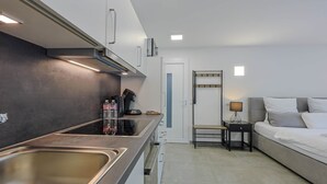 Cozinha compacta com pia, fogão e bancada. Há uma cama com mesa de cabeceira e luminária.