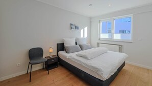 Un dormitorio moderno con cama, una silla, una mesita de noche y un ventanal con persianas.