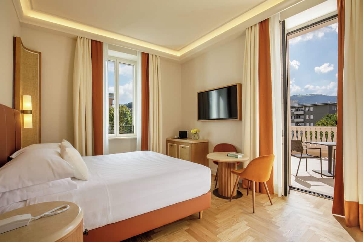 A hotel room with a large bed, a bedside table with a phone, a desk with a chair, a TV mounted on the wall, and a balcony with outdoor seating.
