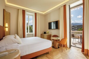 A hotel room with a large bed, a bedside table with a phone, a desk with a chair, a TV mounted on the wall, and a balcony with outdoor seating.