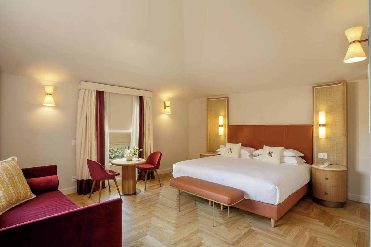 A hotel room with a large bed, a red sofa, a small table with a chair, and a bench.