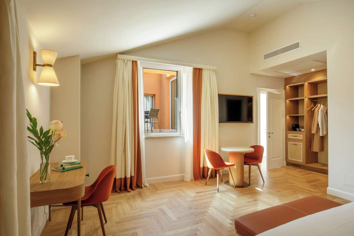 A hotel room with a wooden floor, a dining table with chairs, a TV mounted on the wall, and a walk-in closet with shelves.