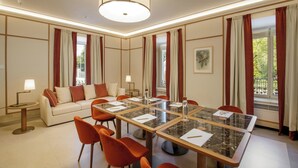 A hotel room with a large marble dining table, orange chairs, a sofa with red cushions, and a large window with curtains. - Villa Miliani (Fabriano)