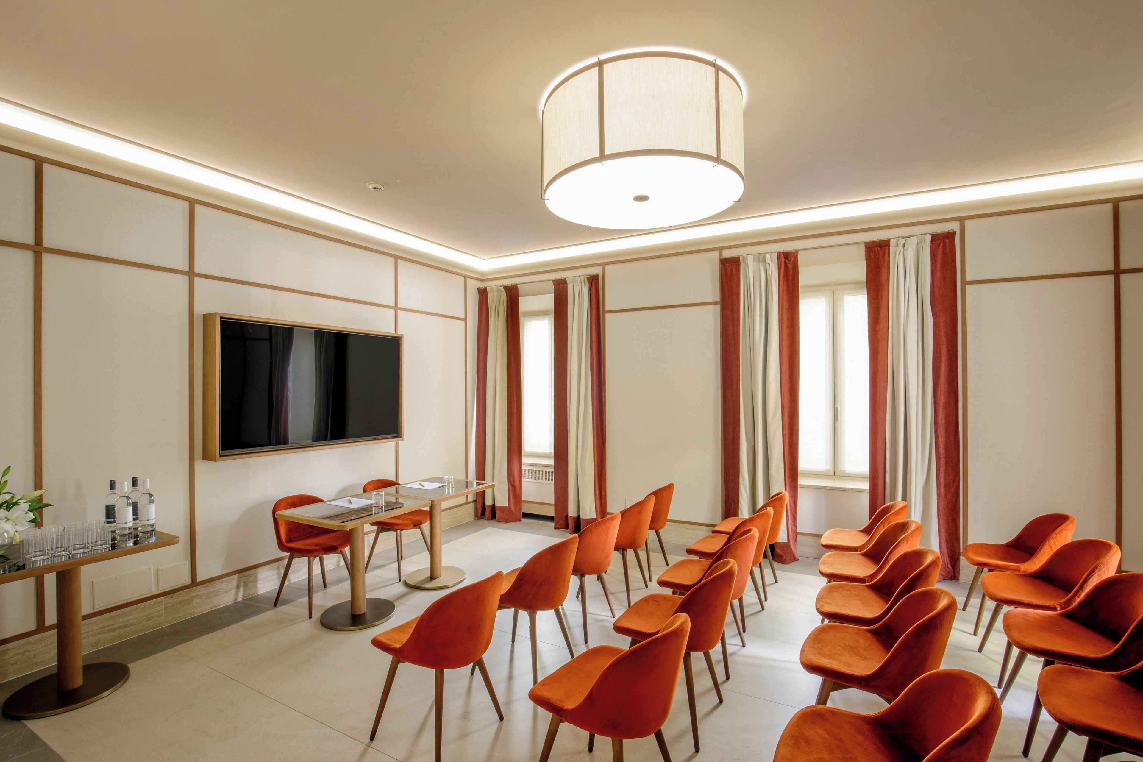 Un salón de conferencias con una amplia pantalla plana, una mesa redonda con sillas y una larga fila de sillas naranjas.