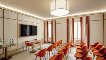 Un salón de conferencias con una amplia pantalla plana, una mesa redonda con sillas y una larga fila de sillas naranjas.