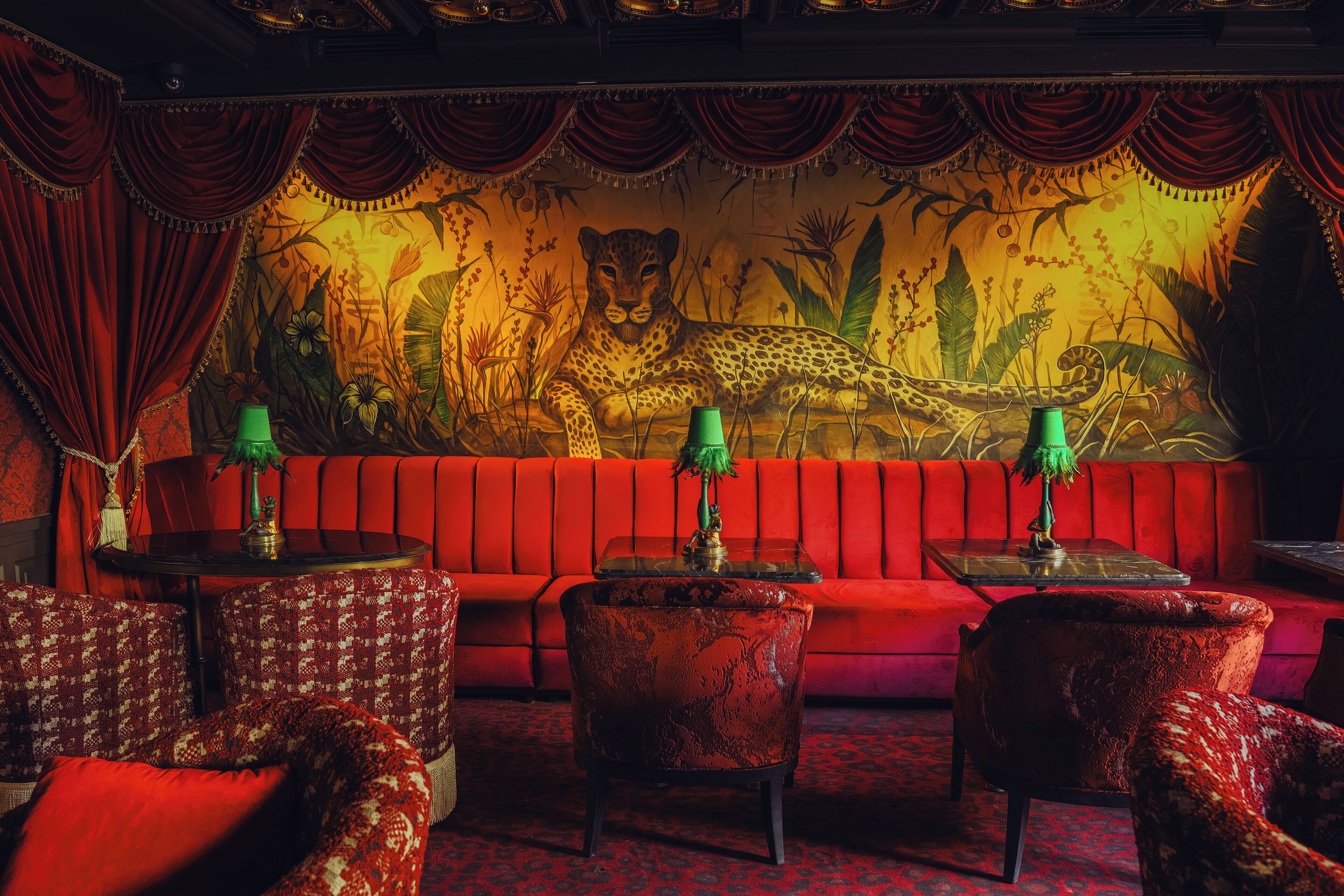 A red sofa set with patterned armchairs, a glass coffee table, and a wall mural of a leopard in a jungle.