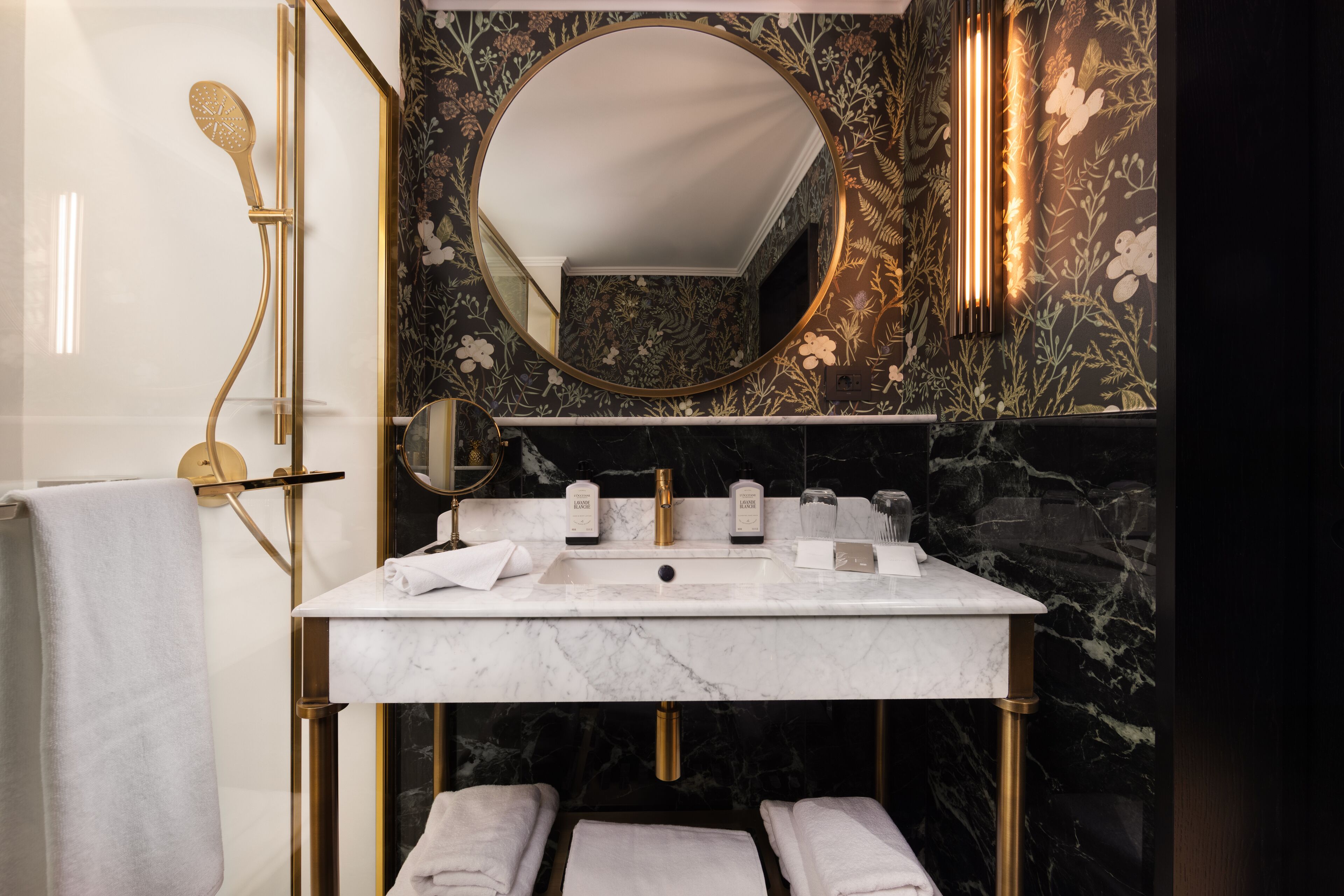 Een badkamer met een marmeren wastafel, gouden details, een grote spiegel en een behang met bloemenprint.