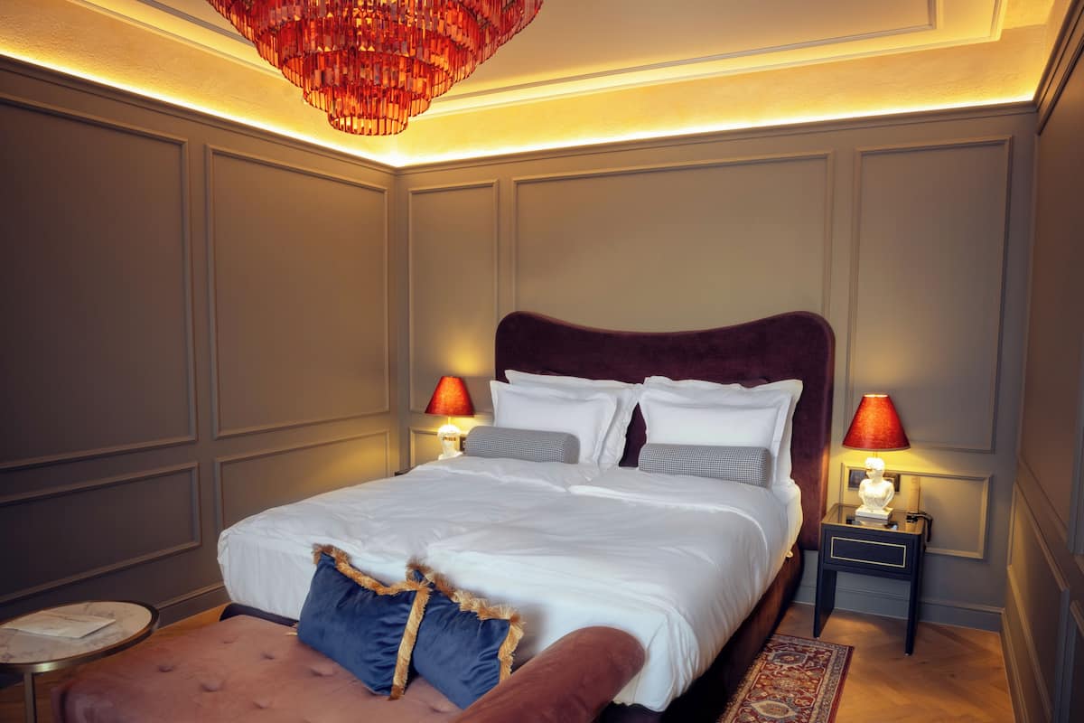 A neatly made bed with white linens and two pillows, a red upholstered bench, and two bedside lamps with red shades.