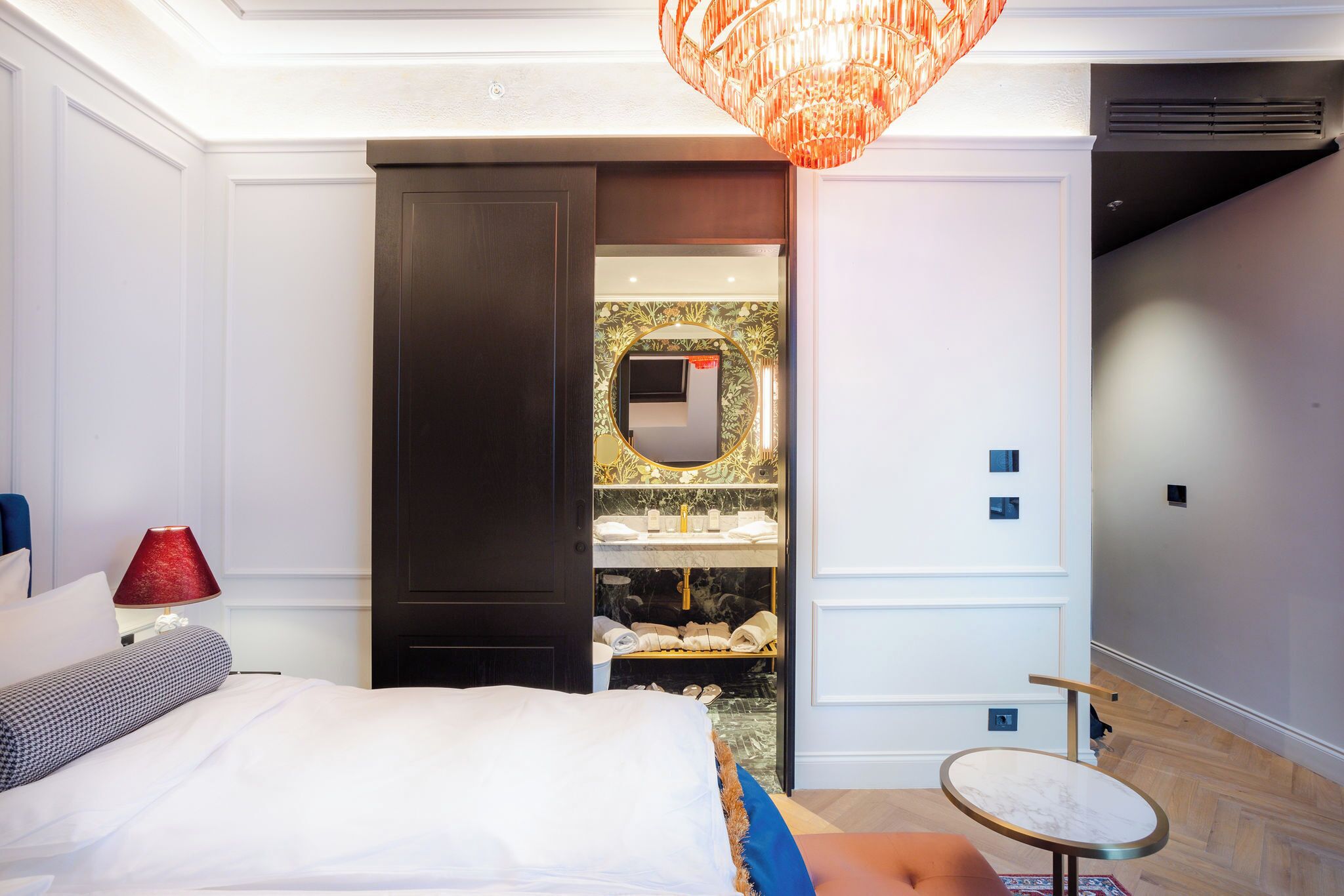 A bedroom with a bed, a nightstand, a red lamp, a round table, and a walk-in closet with a mirror and shelves.