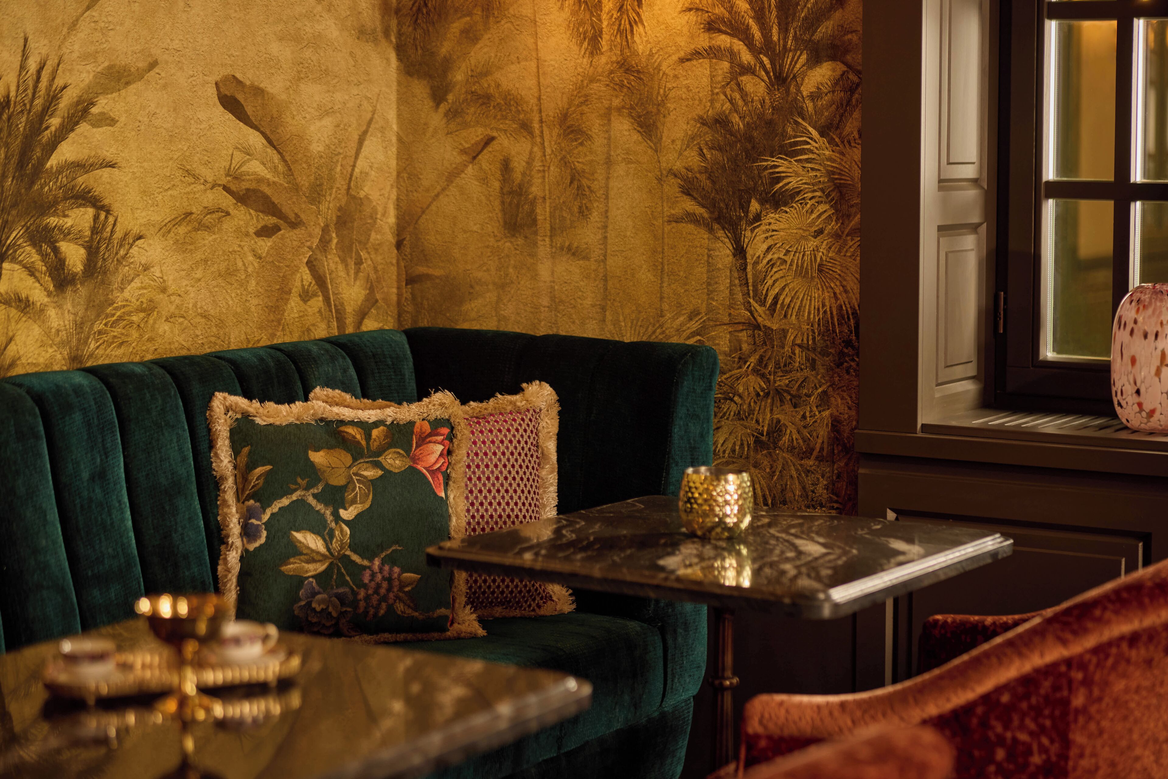 A room with a green velvet sofa, a small table with a glass top, and a window with a view of palm trees.