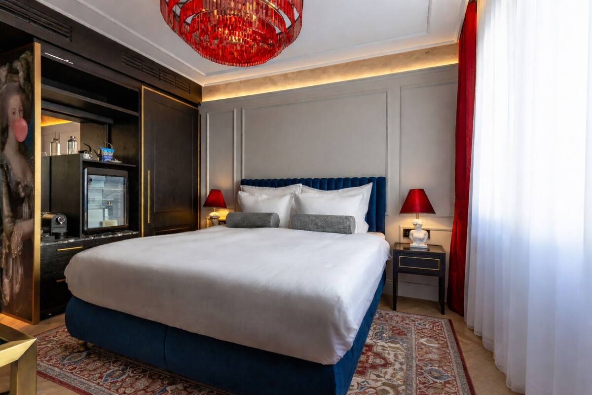 A hotel room with a large bed, two bedside tables with lamps, a red chandelier, and a television on a cabinet.
