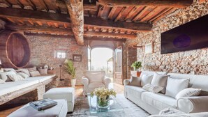 A rustic living room with stone walls, wooden beams, and a large wooden barrel. There are two white sofas, a glass coffee table, and a chair. - Villa Giannello – Tuscan Hillside Hideaway (Matraia)