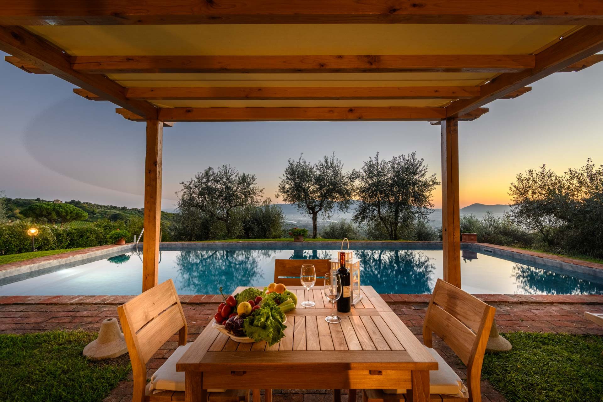 A wooden table with chairs under a pergola, overlooking a swimming pool with a sunset view.