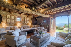 Un intérieur rustique avec des murs en pierre, des poutres en bois et une vue dégagée sur l’extérieur grâce à une porte ouverte.