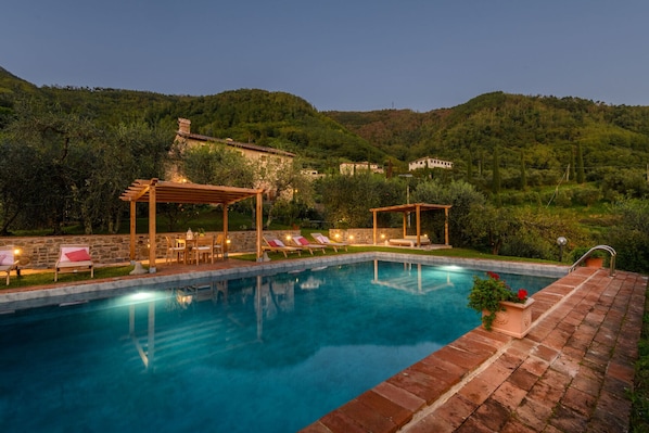 Una piscina con sillones de descanso y una pérgola, rodeada de exuberante vegetación y colinas al fondo.