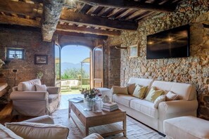 Un intérieur rustique avec des murs en pierre, un plafond en bois, un canapé blanc et une table basse sur laquelle sont posés des livres et un bocal en verre.