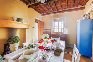 Comedor con mesa dispuesta para cuatro personas, rodeada de sillas. Se observa una ventana con vistas al exterior, un refrigerador azul y un techo de madera.