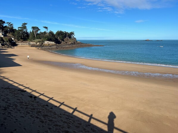 Une plage de sable fin, un ciel bleu dégagé, une personne qui se promène et, au loin, des falaises rocheuses.