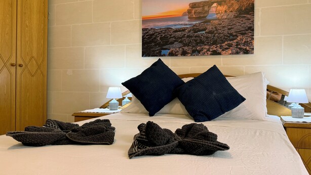 A bedroom with a bed, two nightstands, a wooden wardrobe, and a wall-mounted picture of a coastal landscape. - Traditional Maltese 3BD Apartment (Nadur)