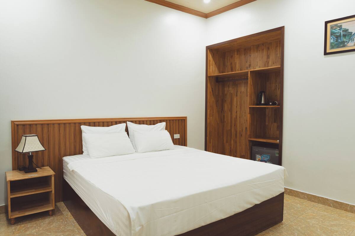 A bedroom with a wooden headboard, a bed with white linens, a nightstand with a lamp, and a built-in wooden shelf unit.