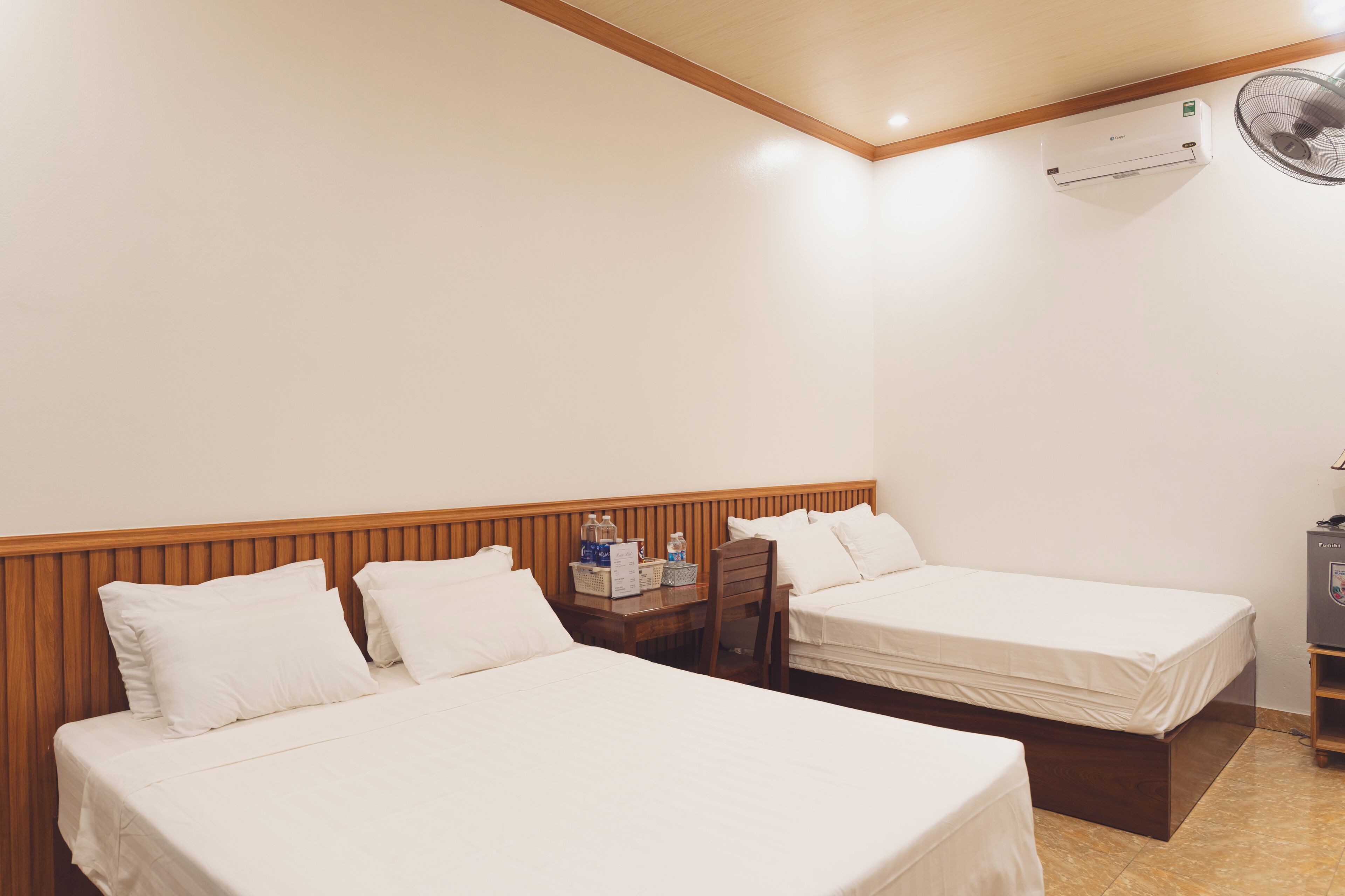 A hotel room with two beds, a wooden headboard, a small table with a fan, and a minibar.