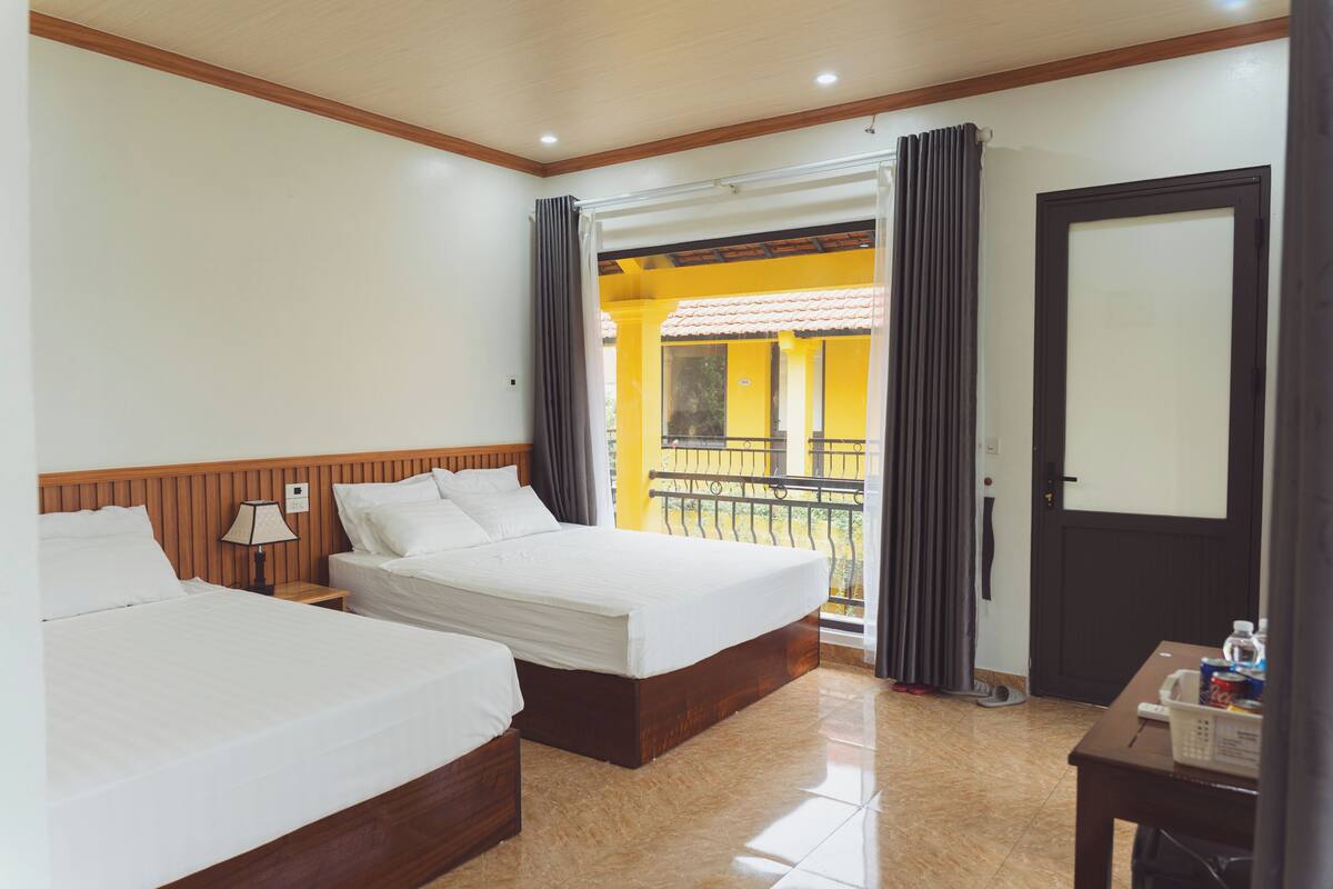 A hotel room with two beds, a small table with a lamp, a door, and a view of the outside balcony.