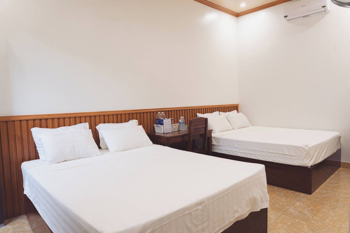A hotel room with two beds, a wooden headboard, a small table with toiletries, and an air conditioning unit on the wall.