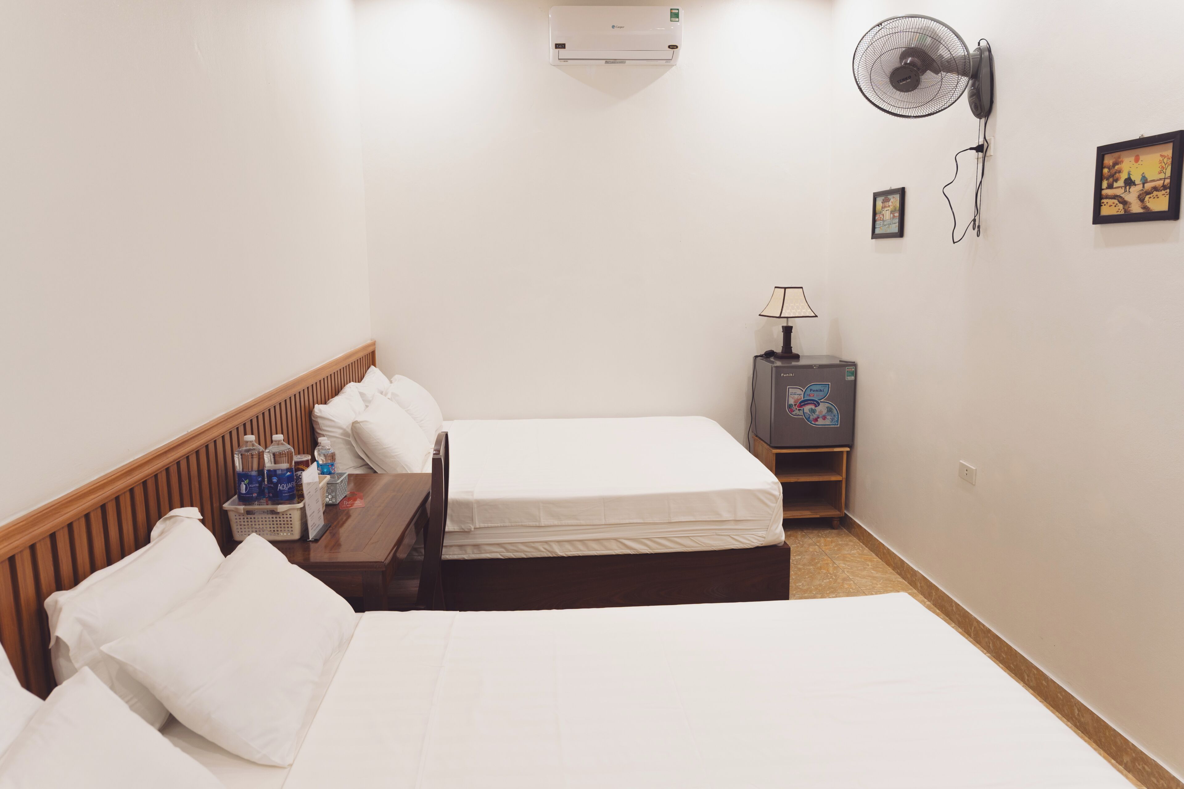Una habitación de hotel con dos camas, una mesita de noche con botellas de agua, una lámpara, aire acondicionado y un ventilador de pared.