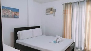 A single-bed room with a dark headboard, white bedding, a small bedside table, a wall-mounted air conditioning unit, and a window with beige curtains. - LIVEWIRE PLANET SUITES (General Santos)