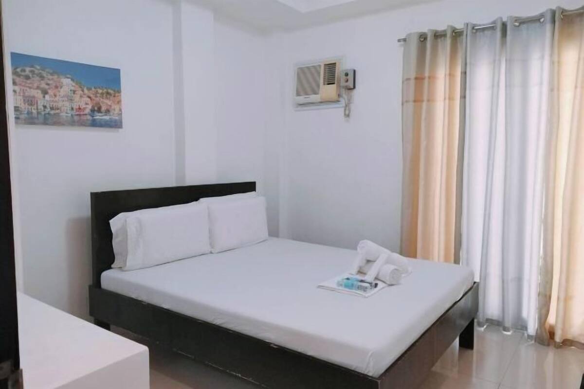 A single-bed room with a dark headboard, white bedding, a small bedside table, a wall-mounted air conditioning unit, and a window with beige curtains.