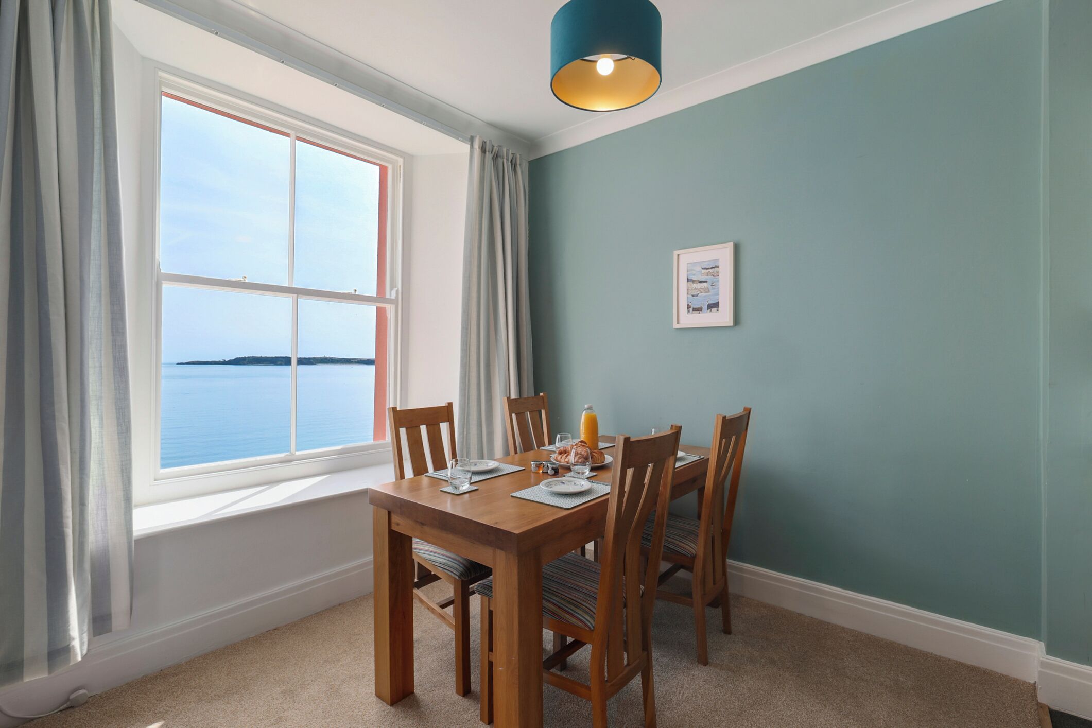 A dining area with a wooden table and chairs, a window with curtains, a framed picture on the wall, and a teal-colored wall.