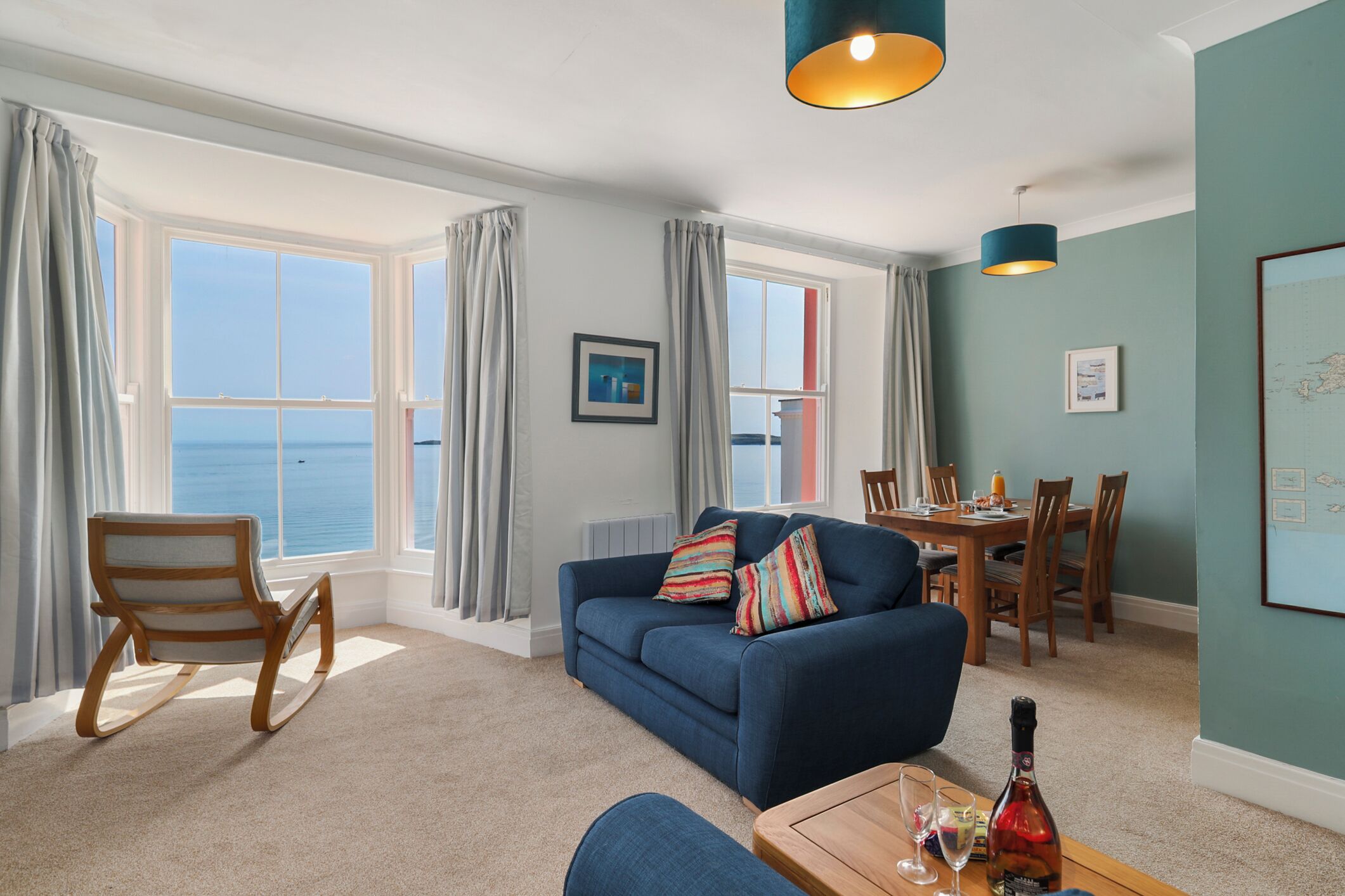 Uma sala de estar com um sofá azul, uma poltrona de balanço de madeira, uma mesa de jantar com cadeiras e vista para o mar através de grandes janelas.