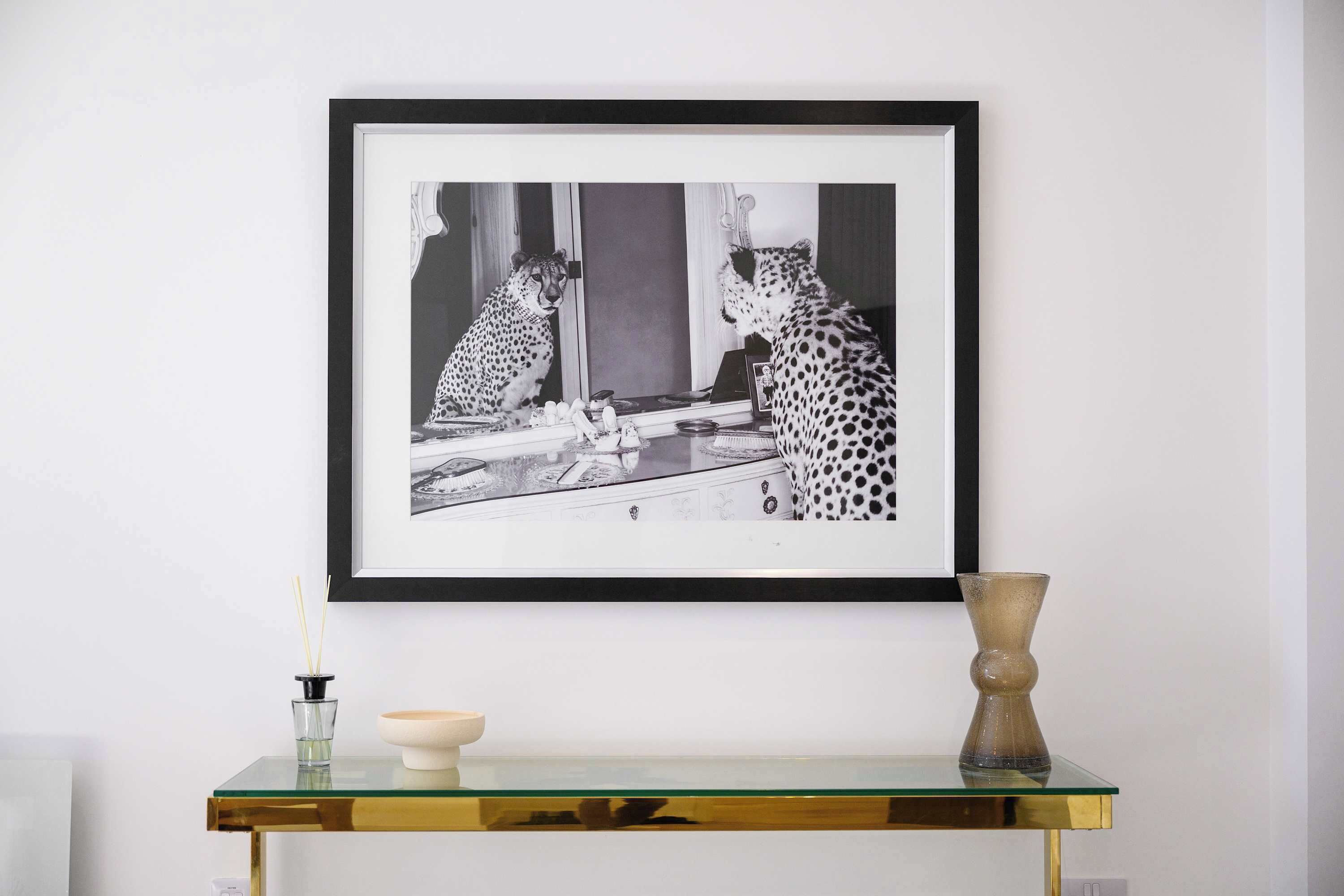 Een zwart-wit foto in een lijst van een cheetah op een glazen tafel met een vaas en een kaars.