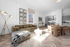 A modern living room with a sofa, coffee table, and floor lamp. There is a dining area with a table and chairs. The room has a large window and a kitchen in the background. - 2 Bed Altrincham Flat | Free Parking (Greater Manchester)