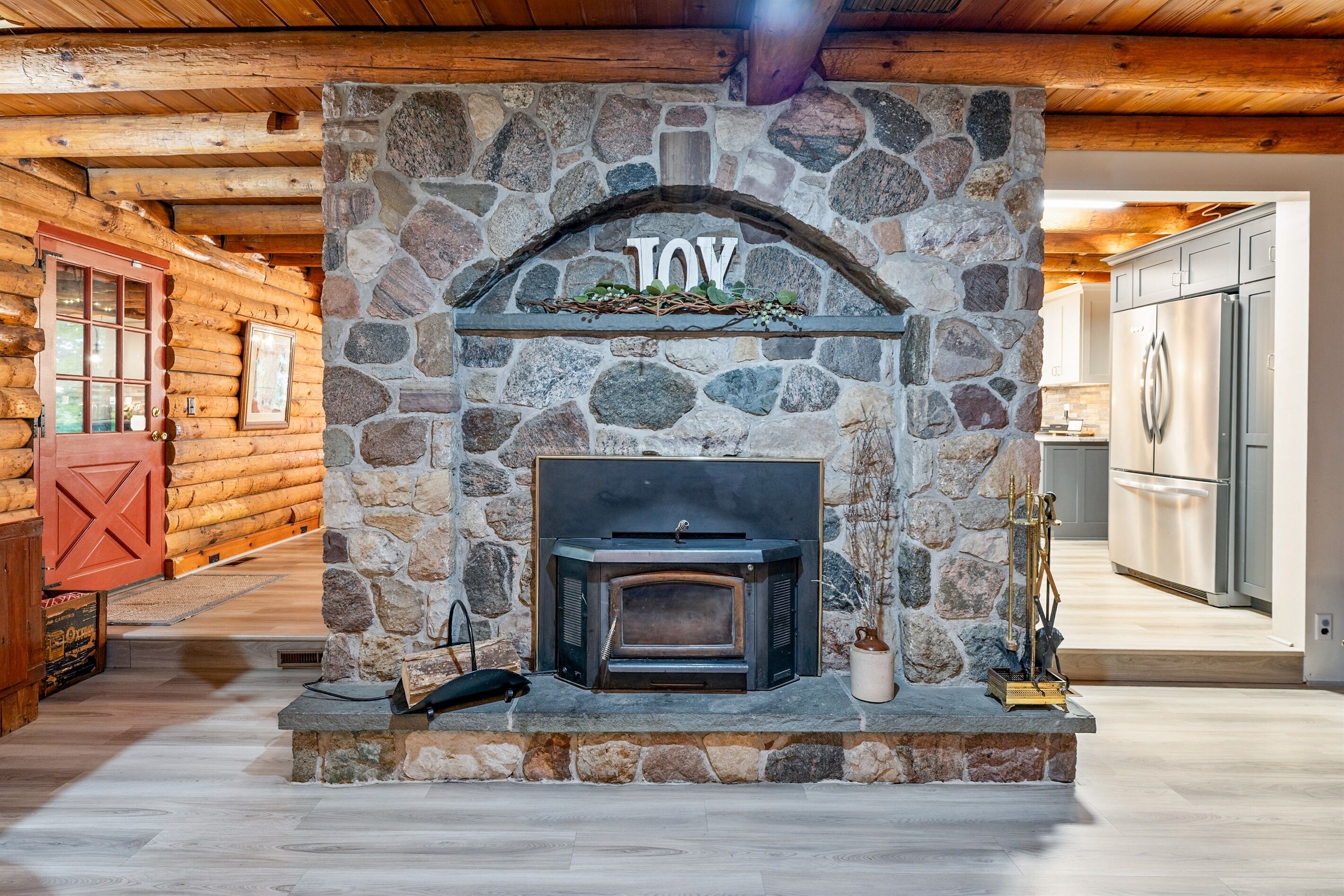A stone fireplace with a "JOY" sign above it, a wooden ceiling, and a kitchen area visible through an open door.