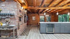 A rustic kitchen with a brick wall, wooden beams, and a large island with gray cabinets. - Canandaigua Cabin Retreat | Lake Views | Hot Tub (Canandaigua)