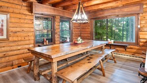 A wooden cabin interior with a large dining table, benches, and large windows offering a forest view. - Canandaigua Cabin Retreat | Lake Views | Hot Tub (Canandaigua)
