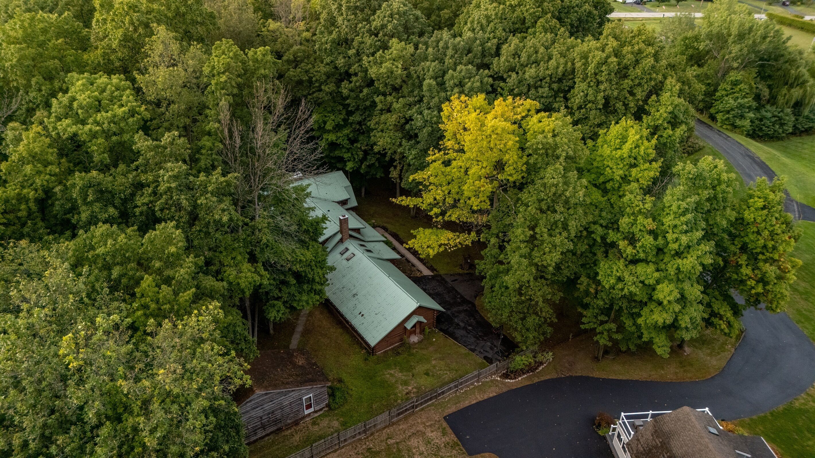 An aerial view of a house surrounded by dense trees, with a visible driveway and a small fenced area.