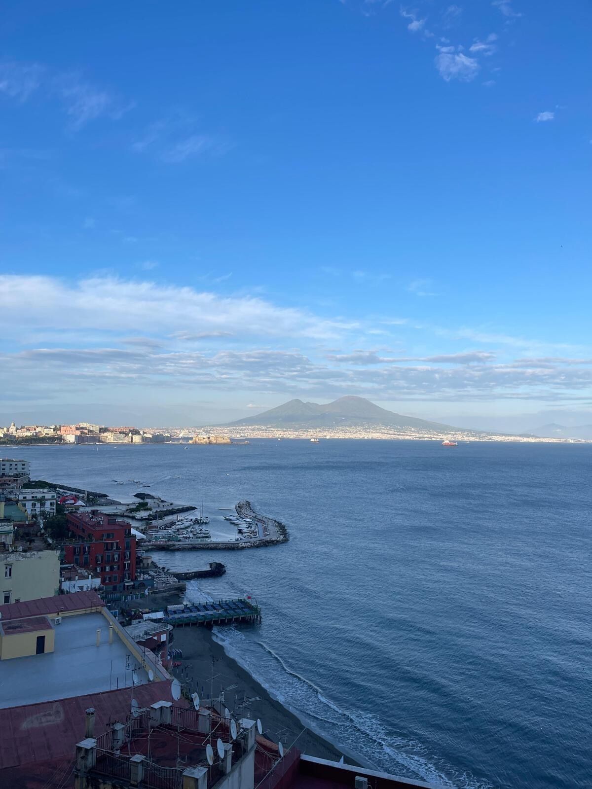 A coastal cityscape with buildings along the waterfront, a pier extending into the sea, and a mountain in the distance.