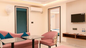 A modern hotel room with a pink sofa, a small table, a floral armchair, a TV mounted on the wall, and a wooden console table with a mirror and a thermostat. - The Maheshwar Hotel (Varanasi)